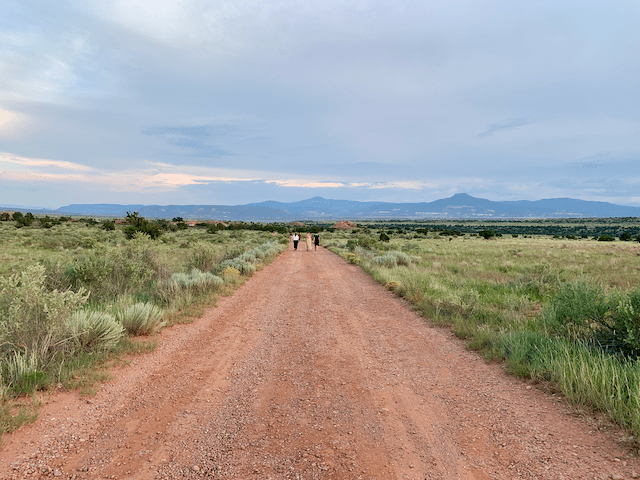 3 people, one in a flowing dress, walking away from the camera on a dirt road in a mountain desert scene